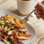 Apple Carrot Salad served on a white plate with a hand holding a fork, featuring carrots, apples, walnuts, and orange slices, accompanied by a cup of herbal tea on a marble countertop