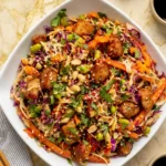 Overhead view of Asian Chicken Crunch Salad topped with cilantro, sesame seeds, and peanuts, served with wooden chopsticks, soy sauce, and chili oil on a marble countertop
