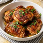 Baked Chicken Thighs served in a white bowl, golden-brown with herb garnish, placed on a striped cloth over a marble countertop