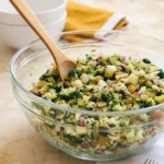 Broccoli Salad in a clear glass bowl with a wooden spoon, featuring broccoli florets, apples, almonds, and dried cranberries, served on a marble countertop with white bowls and a yellow napkin