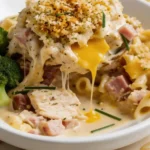 Chicken Cordon Bleu Casserole in a white bowl, melted cheese stretching over chicken, ham, pasta, broccoli, and fresh chive garnish