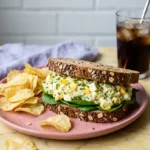 Egg Salad Sandwich served on whole grain bread with potato chips and a glass of iced beverage on a pink plate over a marble countertop
