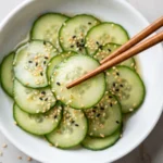 Japanese cucumber salad in white bowl — thinly sliced cucumbers with sesame seeds and light dressing, lifted by wooden chopsticks