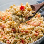 Close-up of macaroni salad being spooned, featuring Macaroni Salad, red bell peppers, celery, and creamy cheese dressing, with a wooden spoon