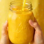 Fresh Mango Smoothie in a glass jar with a yellow straw, held by hands against a bright yellow background