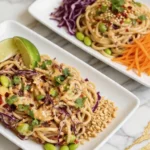 Thai Noodle Salad served on white rectangular plates featuring noodles, purple cabbage, shredded carrots, edamame, lime wedge, peanuts, and fresh herbs on a marble countertop with gold accents