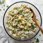 Tuna pasta salad in glass bowl with shell pasta, peas, celery, dill, and creamy dressing - overhead view on marble countertop with wooden serving spoon