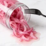 Pickled red onions in a glass jar with a fork, vibrant pink rings spilling onto a white marble countertop
