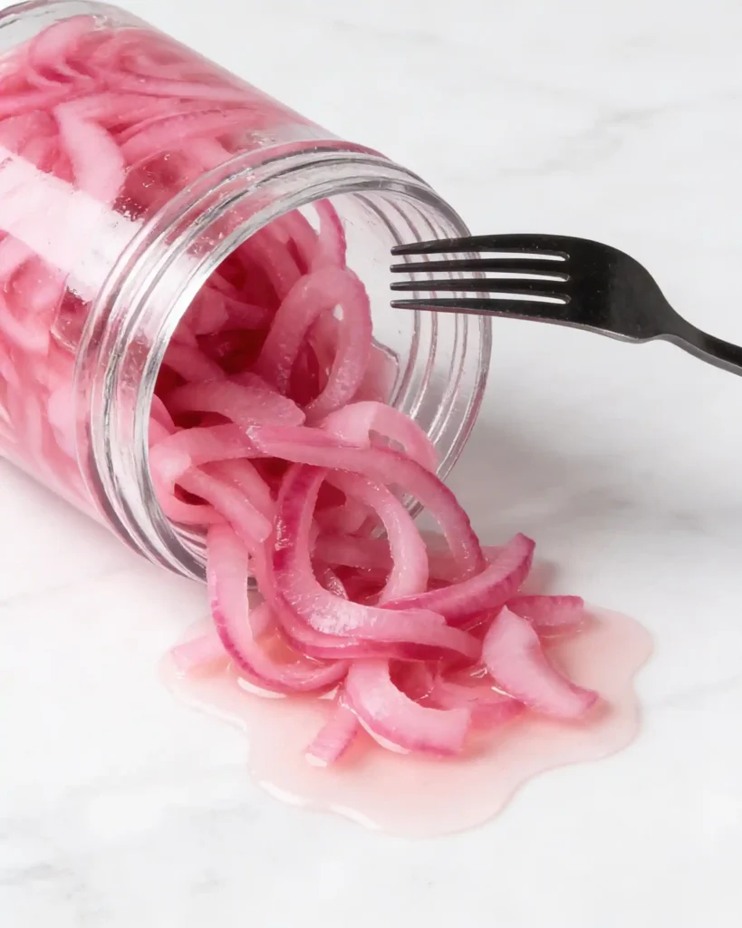 Pickled red onions in a glass jar with a fork, vibrant pink rings spilling onto a white marble countertop