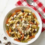 Overhead view of Chicken Salad Chick inspired pasta salad in white bowl. Features tri-color rotini, artichoke hearts, black olives, crumbled feta and grated Parmesan. Served beside red-and-white checkered napkin with spoon