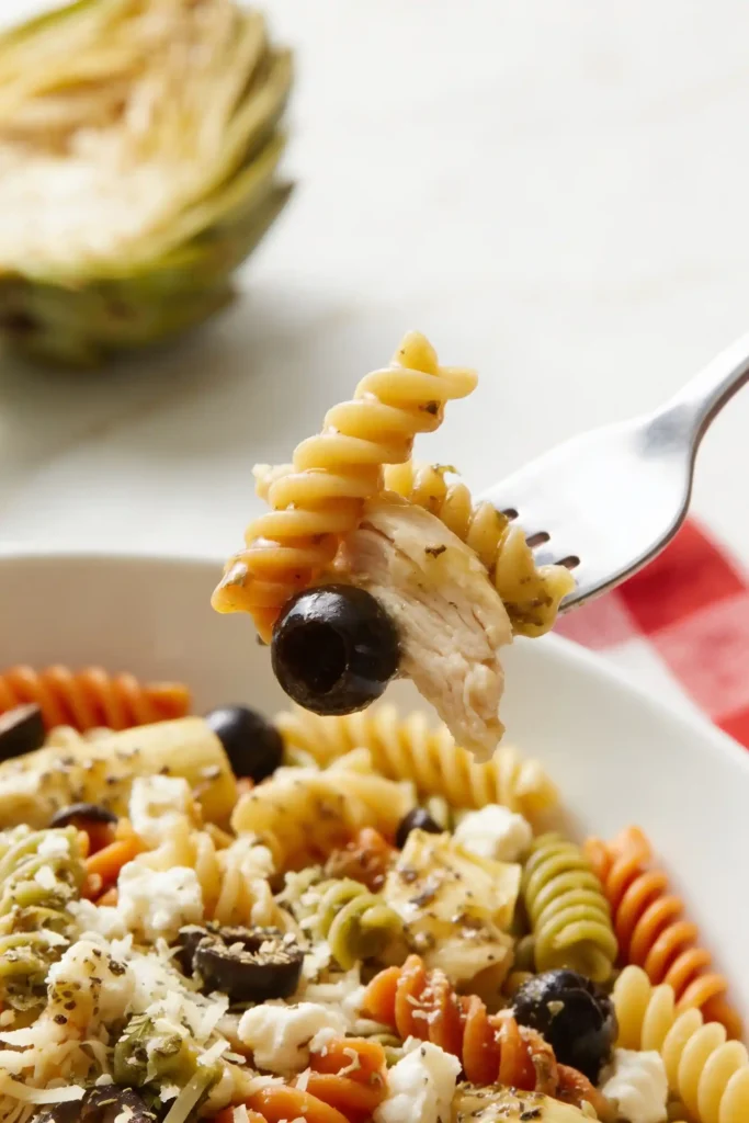 Close-up of fork holding rotini pasta, shredded chicken and black olive from Chicken Salad Chick style pasta salad. Background shows bowl with artichoke hearts, feta, olives and tri-color noodles
