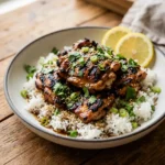 Bowl of Hawaiian chicken over fluffy coconut rice, topped with cilantro, scallions, sesame seeds. Lemon wedges on side. Rustic wood background