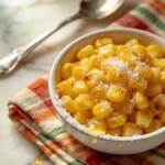 Close-up of KFC-style sweet corn in ceramic bowl topped with coarse sea salt, cracked pepper and glistening honey drizzle. Bowl rests on folded plaid napkin beside vintage silver spoon on gold-veined marble.