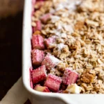 Unbaked rhubarb crisp in white ceramic baking dish. Chopped pink rhubarb topped with brown sugar, oats, butter cubes, and coarse sugar. Dish rests on folded linen napkin with red stripe over dark wood surface. Ready for oven