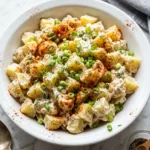 Creamy potato salad in white bowl topped with chives, dill, paprika. Spoon and peppercorns beside it on marble. Classic side dish