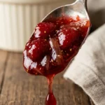 Close-up of spoon holding thick, glistening strawberry rhubarb jam with visible fruit pieces and seeds. A drop falls from the spoon onto rustic wood surface. Background shows blurred ramekin and cloth napkin