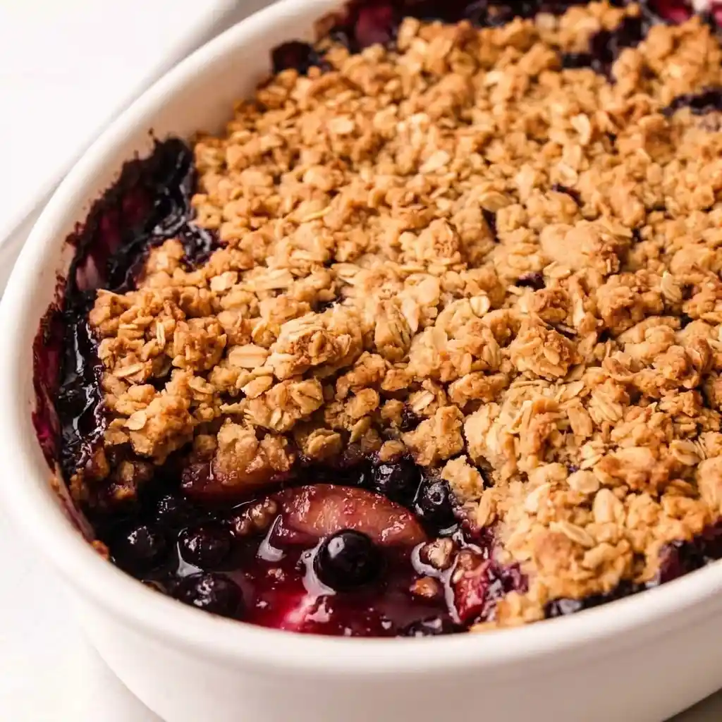 Apple blueberry crisp in white baking dish with scoop removed to show juicy fruit filling under oats